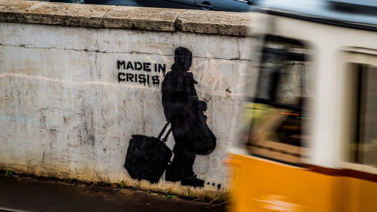 Street art with a stencil of a person walking with bags and the words "Made in Crisis."