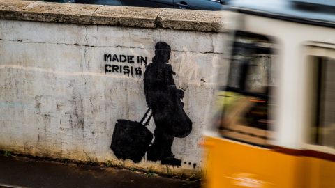 Street art with a stencil of a person walking with bags and the words "Made in Crisis."