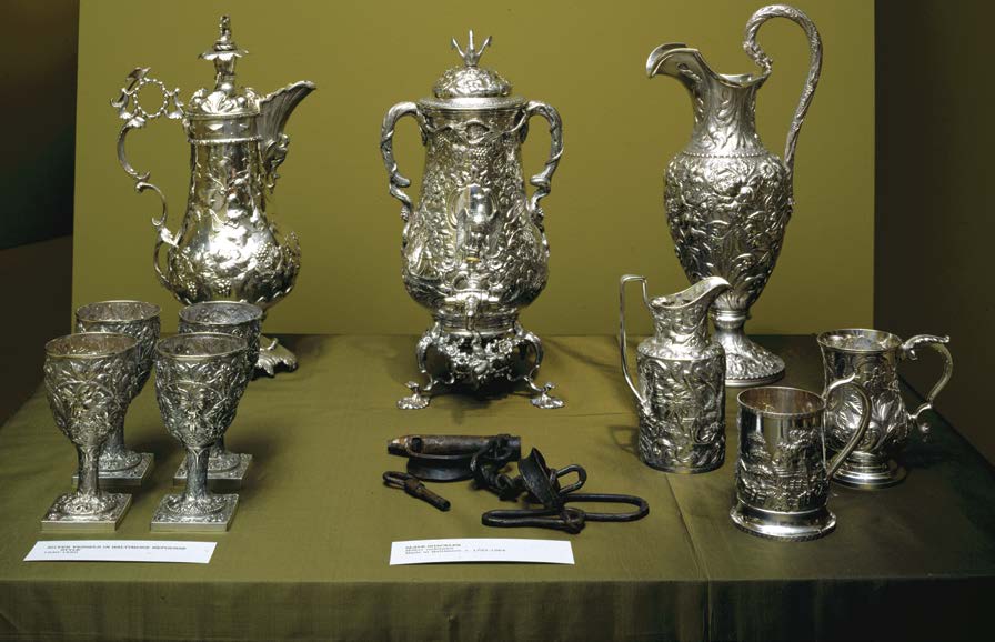 A group of silver teapots and service with a pair of iron shackles displayed between them. 