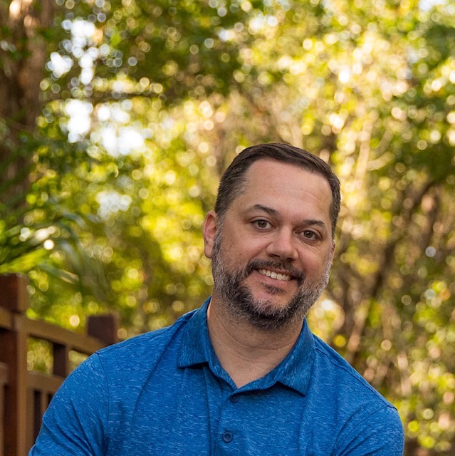 Bearded male in blue polo shirt with trees in the background