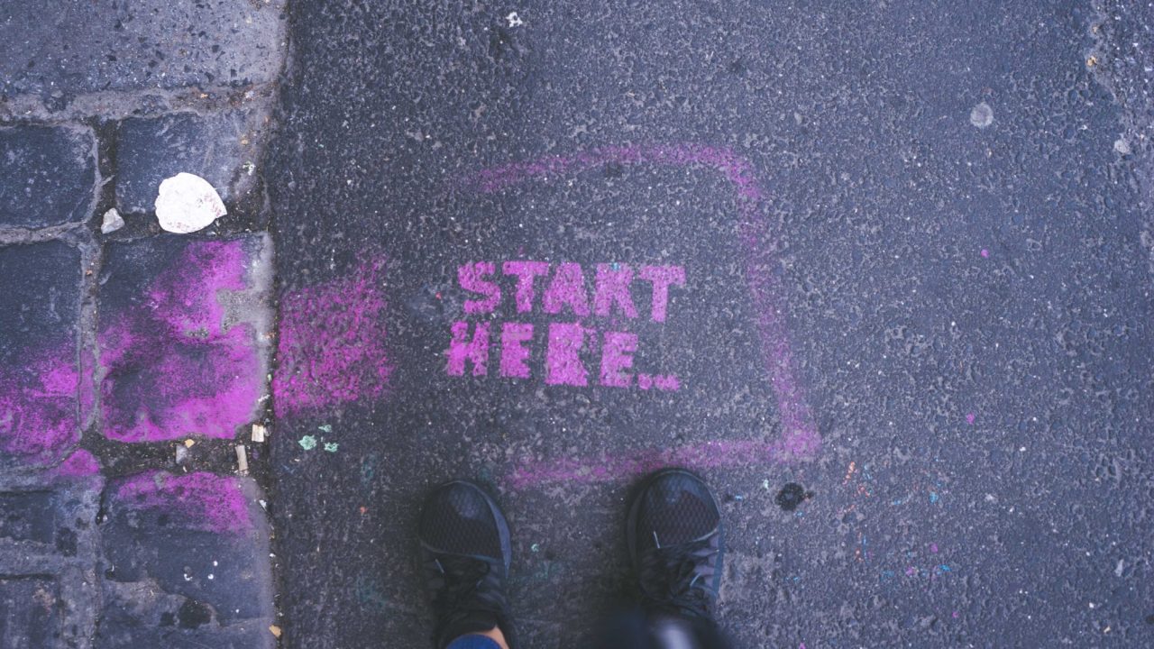 Sneaker-wearing feet standing over sidewalk art reading "Start here..."