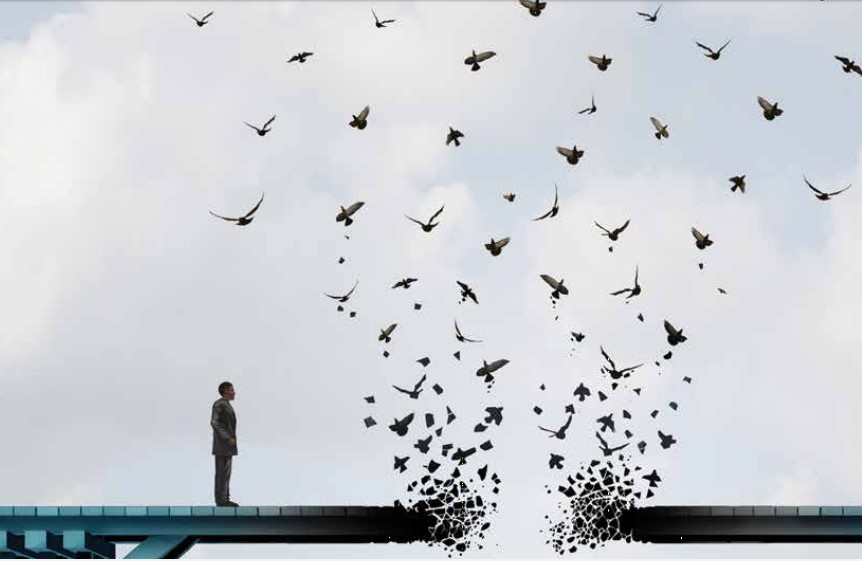 A man stands on a wooden bridge with a break in the middle where dozens of birds are flying off into space.
