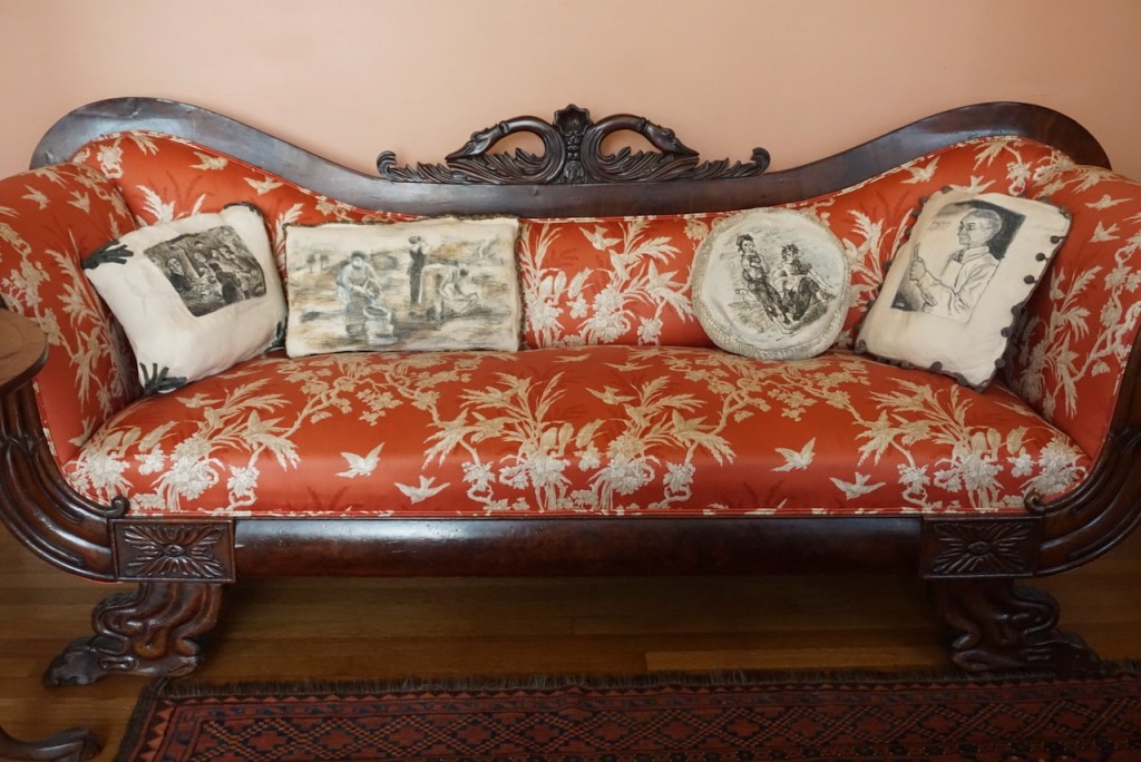 An antique, ornately detailed sofa with decorative pillows showing scenes of 19th-century life.