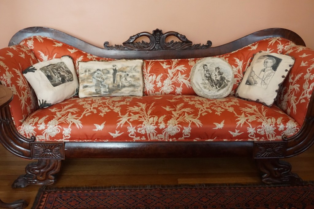 An antique, ornately detailed sofa with decorative pillows showing scenes of 19th-century life.
