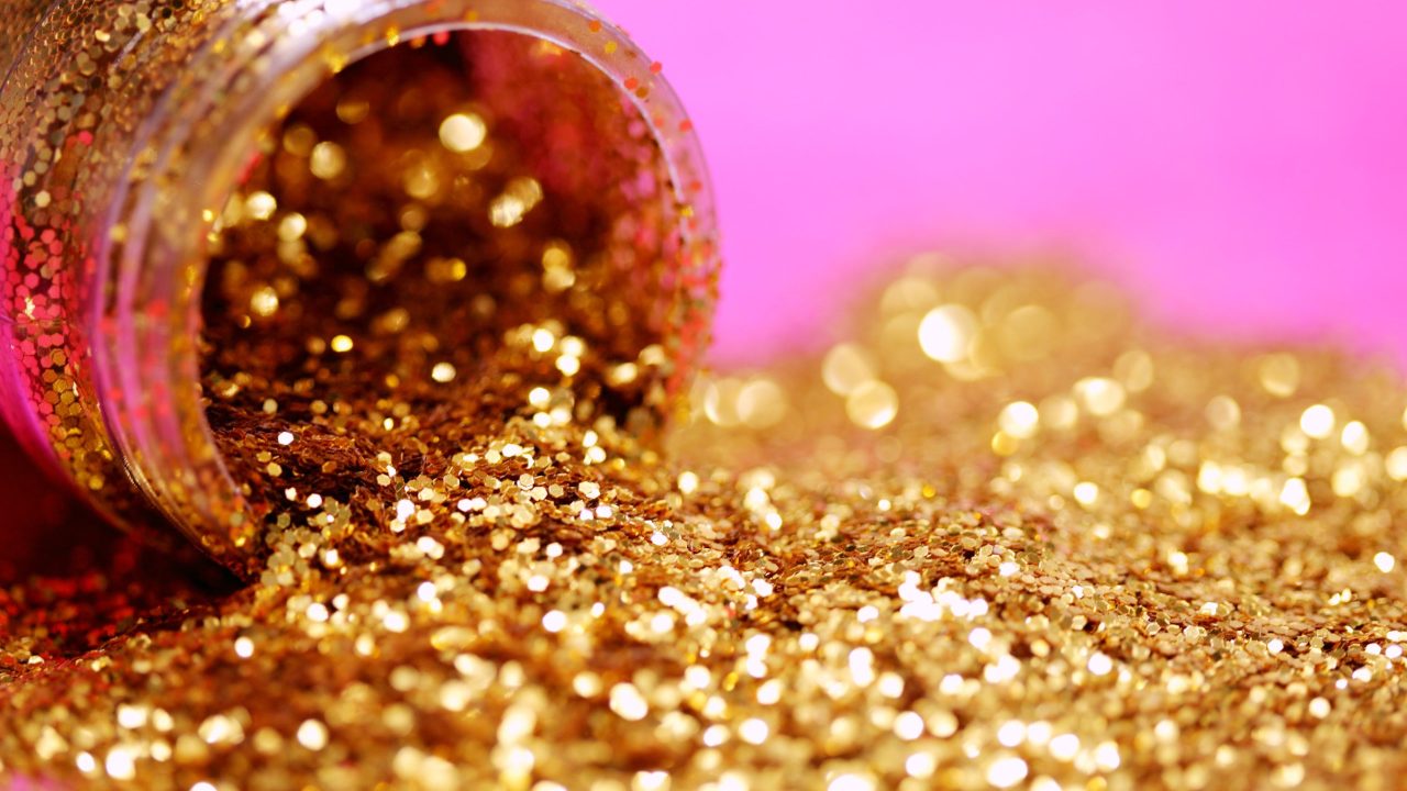 A bottle of gold glitter poured out against a pink background.