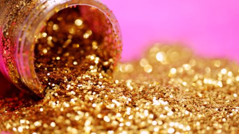 A bottle of gold glitter poured out against a pink background.