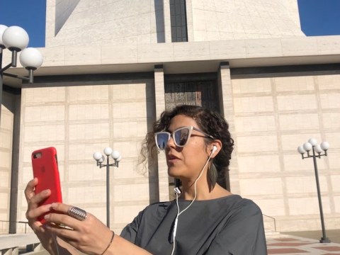 Person standing in front of a building holding up their phone with headphones plugged into it