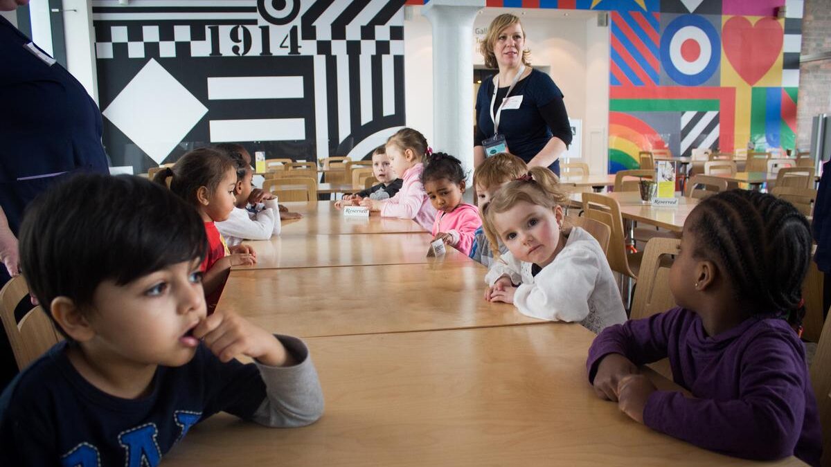 Children sitting around tables in front of graphic wall murals, with a teacher standing in the background