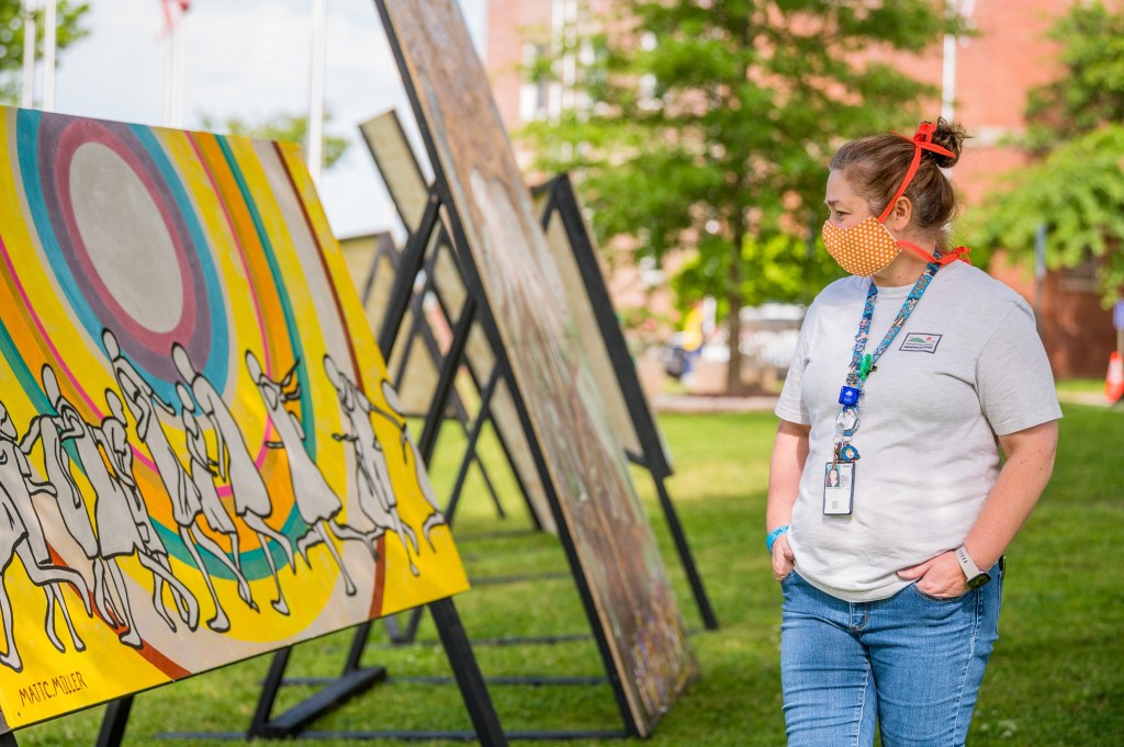 A person looking at the paintings on display outside while wearing a mask and identification badge on a lanyard