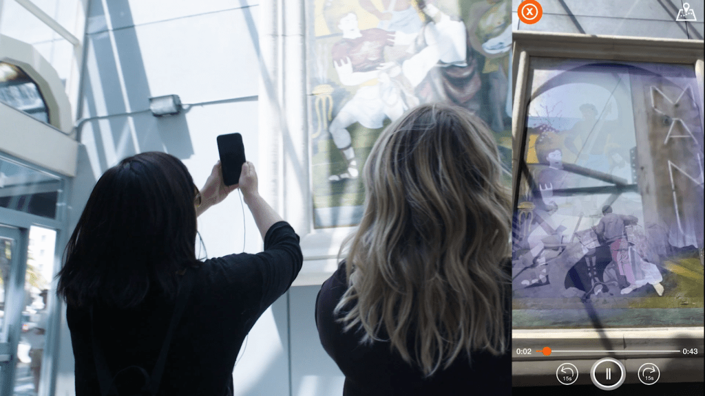 Two people holding their phones up to a mural on a wall, with an image of their screen showing archival footage superimposed over the mural
