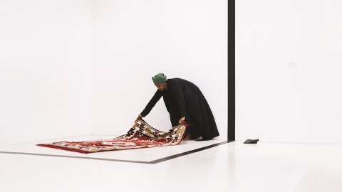 A person lays a prayer mat down in a white walled space.