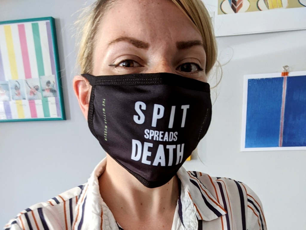 A person posing in from of hanging prints wearing a black mask reading "Spit Spreads Death" in white lettering, with the name of the museum printed on the side.