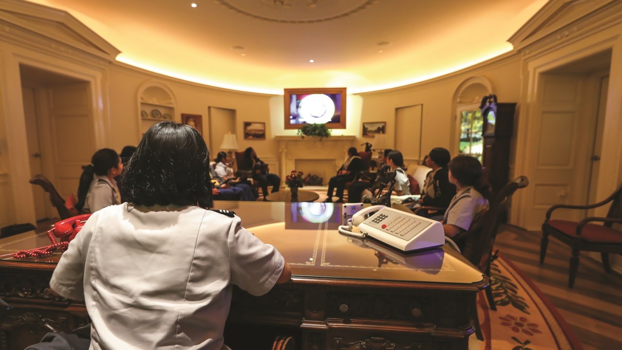A student sits at a desk facing the replica oval office with other students.