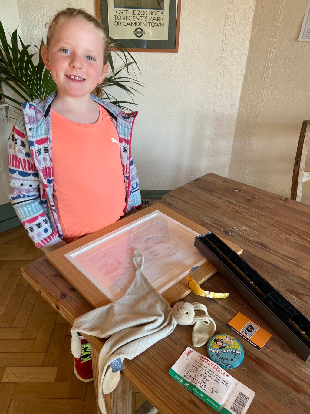 A child poses with an array of objects displayed on a table, including a stuffed animal, a framed drawing, a watch, and a ticket to a sporting event