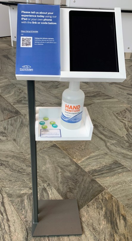 A stand with a sign reading "Please tell us about your experience today using our iPad or your own phone with the link at code below, with a QR code on the sign, an iPad resting on the stand, a bottle of hand sanitizer, and takeaway buttons.