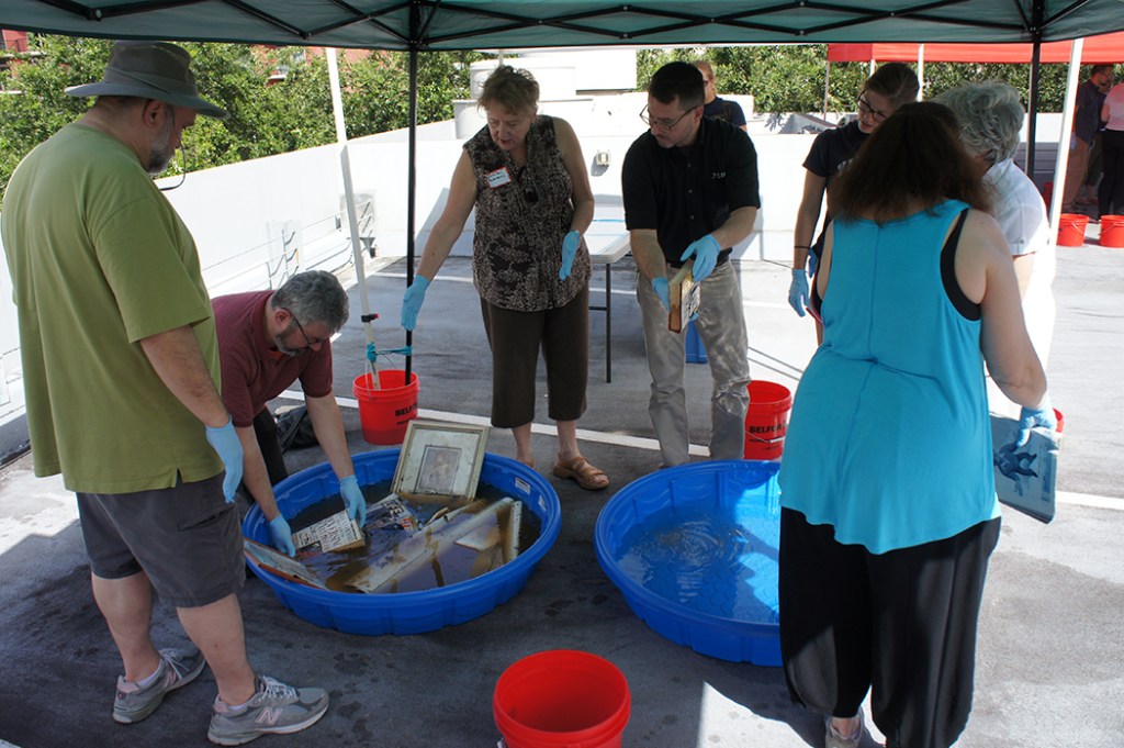 A group of people standing under a tent with buckets and two large plastic saucers under it. One of the saucers is filled with brown water and objects like framed pictures and pieces of paper.