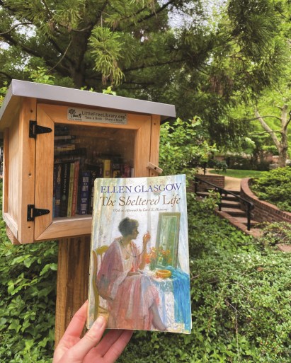 Someone holds a copy of Ellen Glasgow's "The Sheltered Life" in front of a small wooden structure with a roof and clear door on a wooden pole. 