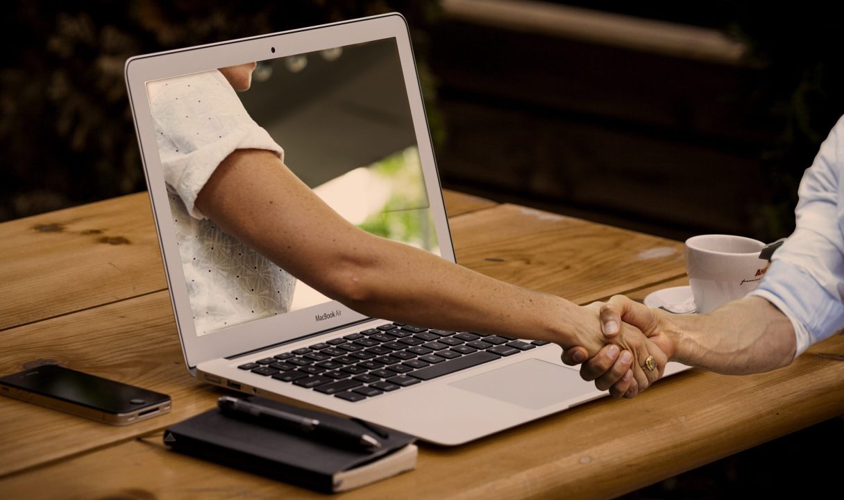 A surreal graphic of a person on a computer screen extending their hand out for a handshake with another person in front of the computer