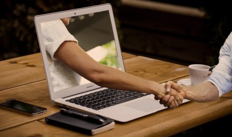 A surreal graphic of a person on a computer screen extending their hand out for a handshake with another person in front of the computer