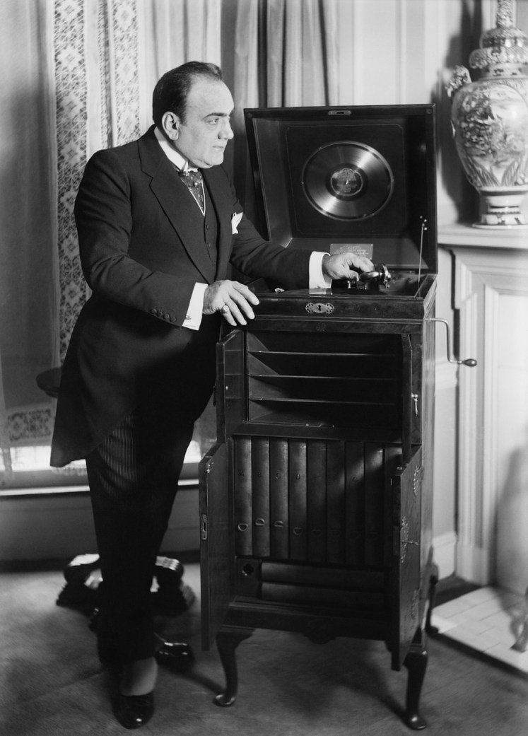 A black-and-white photograph of someone in formal dress operating a Victrola