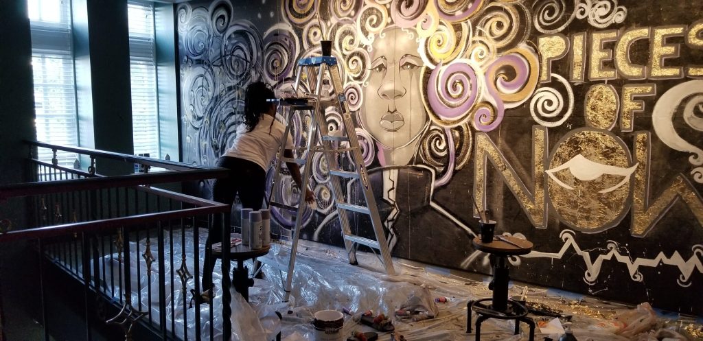 An artist painting a mural indoors of a figure with an abstract swirl of hair and the title "Pieces of Now."