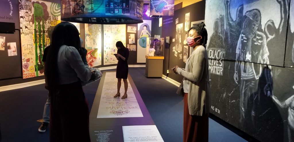 A news reporting team standing inside of an exhibition showing murals hung on walls