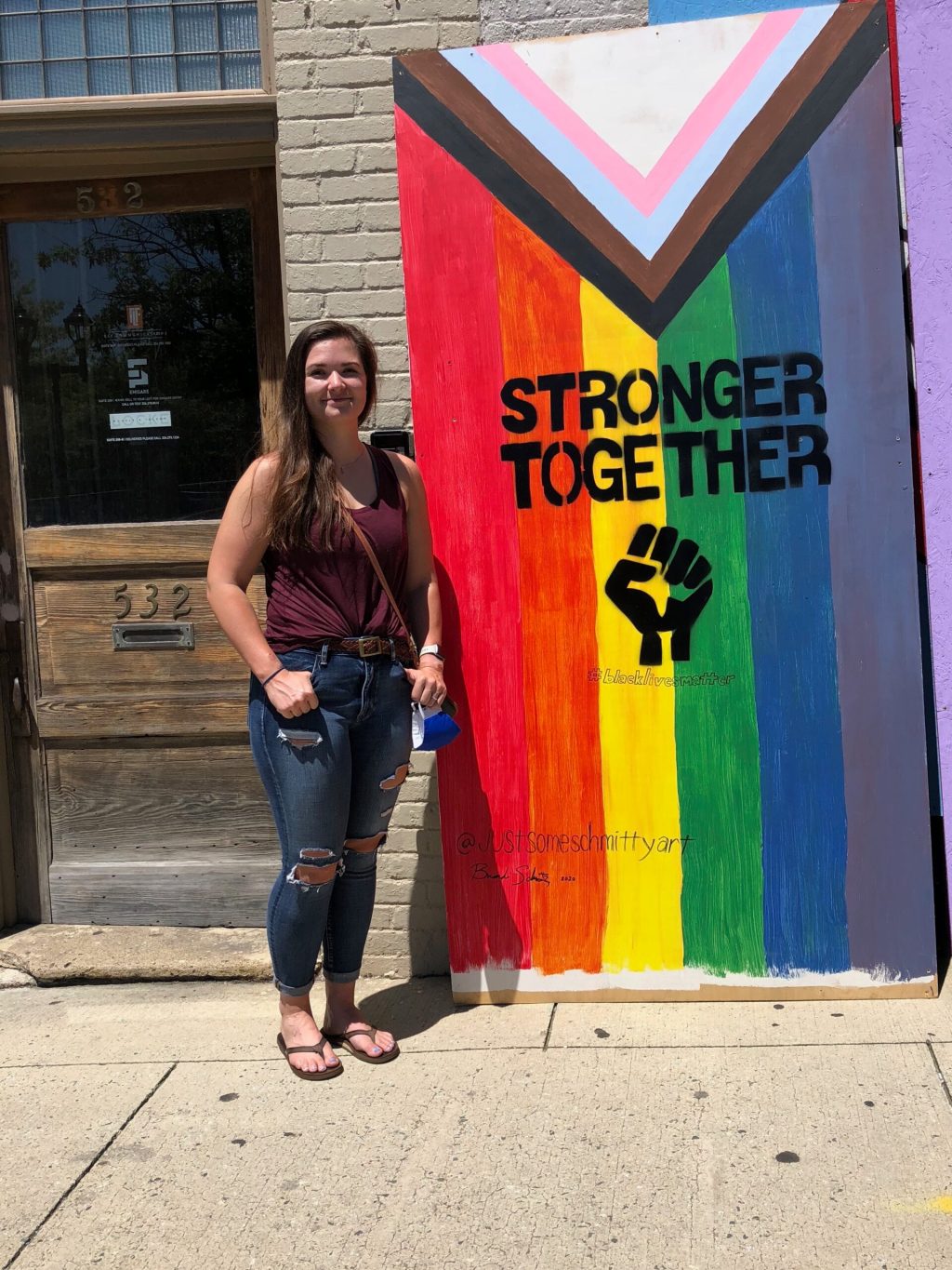 A person standing in front of a mural reading "Stronger Together" with a Black power fist against an inclusive Pride flag backdrop