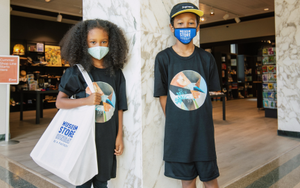 Children posing in a museum space wearing "Museum Store Sunday" branded tote bags and face masks.