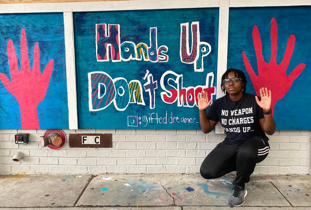 A person wearing a t-shirt reading "No Weapon / No Charges / Hands Up..." in front of a mural reading "Hands Up Dont Shoot" with drawings of hands