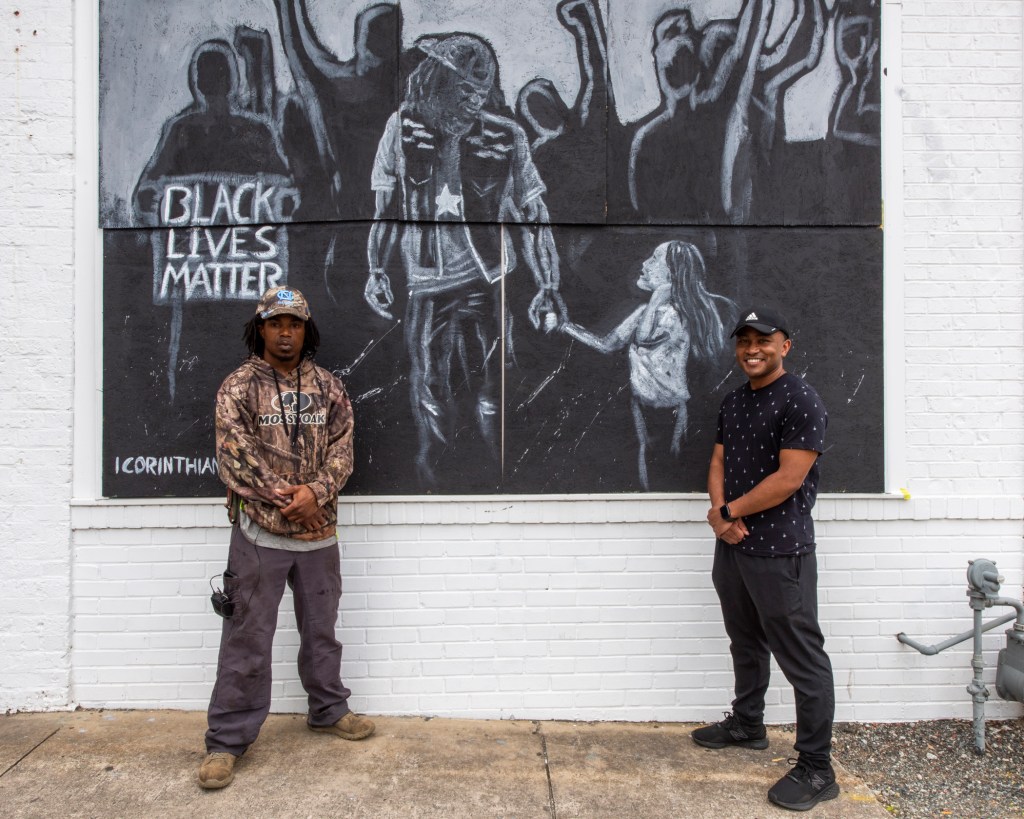 Two people standing in front of a mural depicting a Black Lives Matter protest