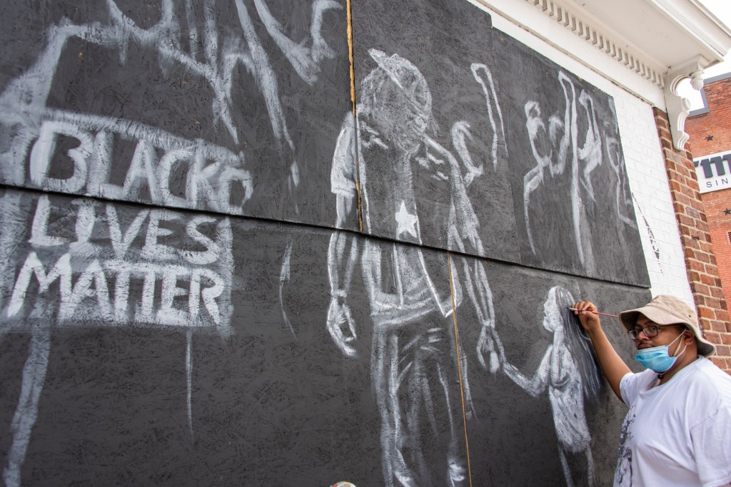 An artist holding a paintbrush working on a mural depicting a Black Lives Matter protest in a loose, minimal style