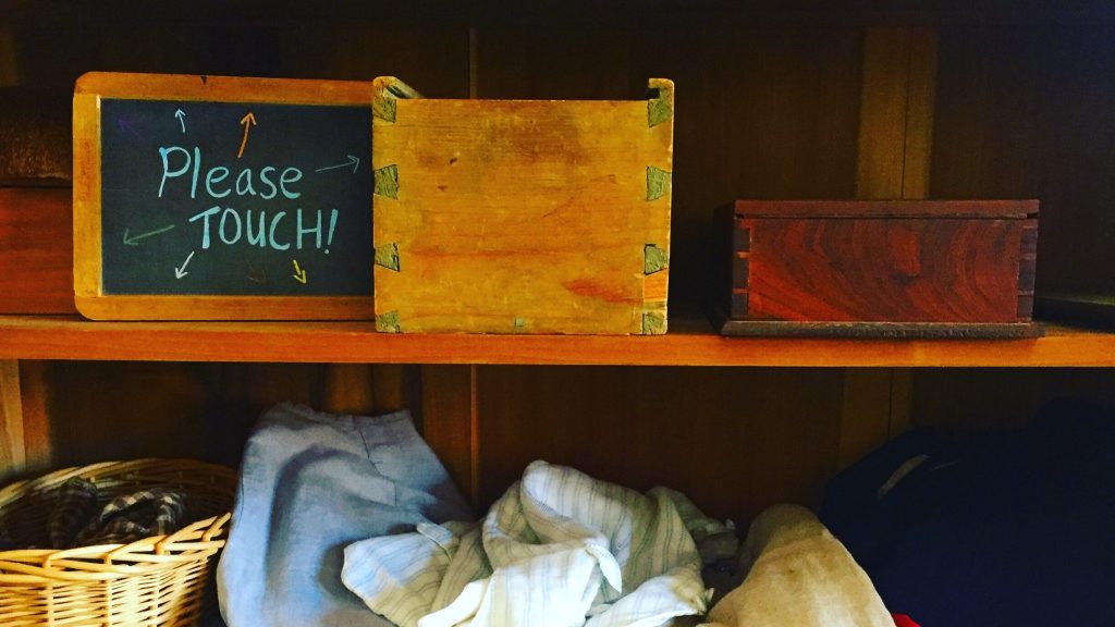 A shelf with period costumes and objects displayed with a chalkboard sign reading "Please Touch!"