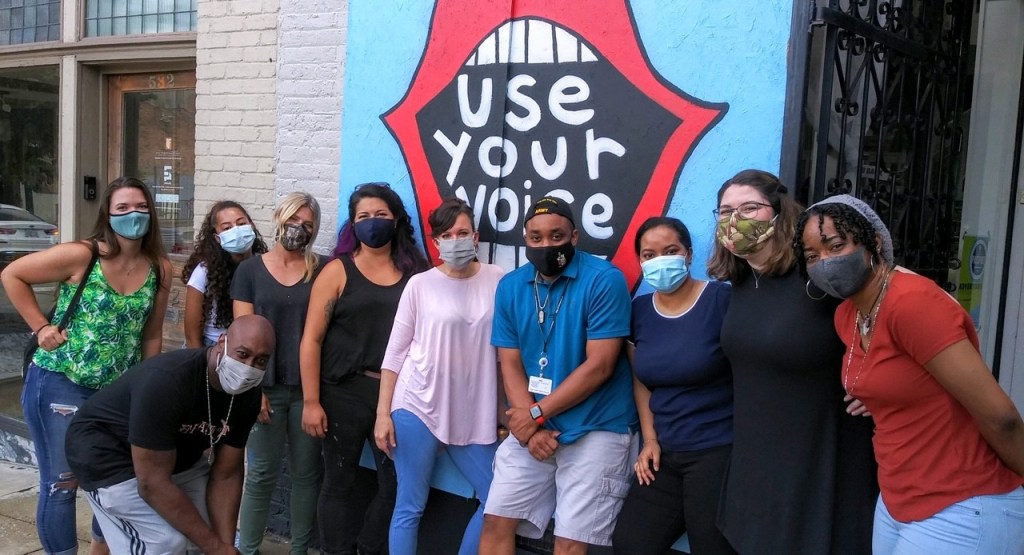 A group of people posing in front of a mural depicting an open mouth with the message "Use Your Voice."