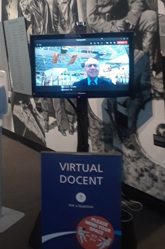 A monitor on a stand reading "Virtual docent: ask a question" with a video feed of a person on screen