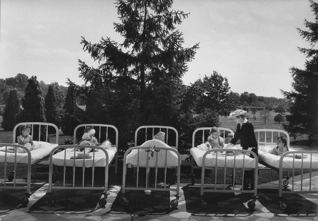 A black-and-white photograph of a woman interacting with children lying in wheeling hospital beds outside