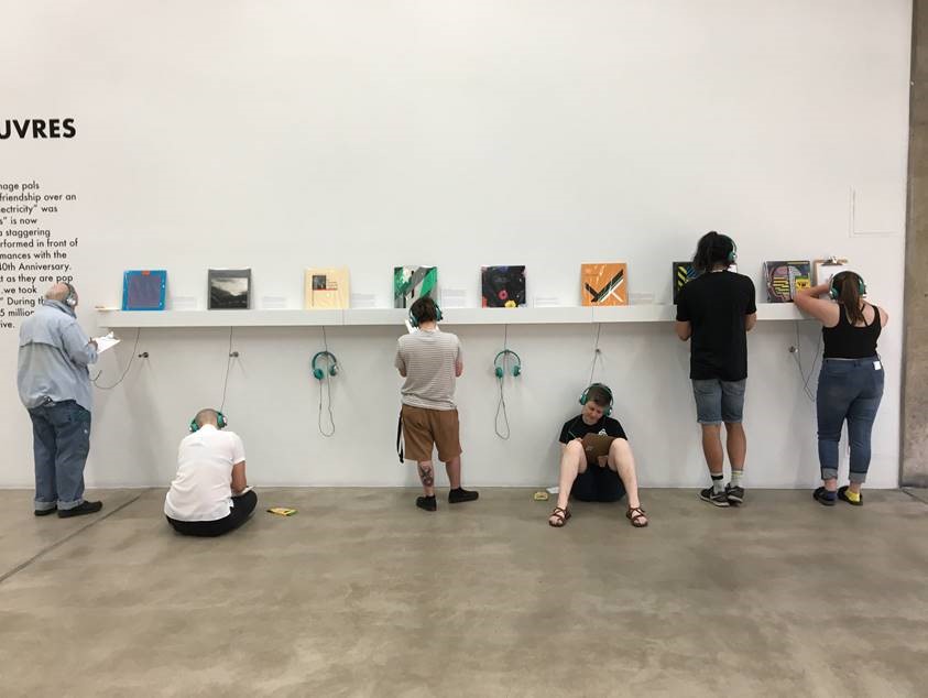 A group of people standing at listening stations in a museum gallery listening and taking notes