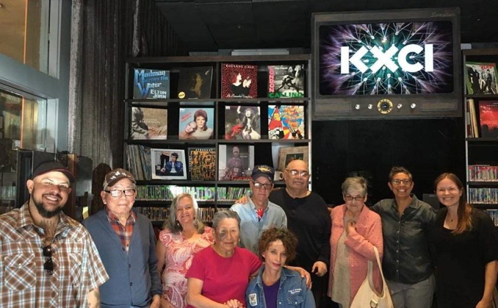 A group portrait in front of a sign that says "KXCI" and a wall of records