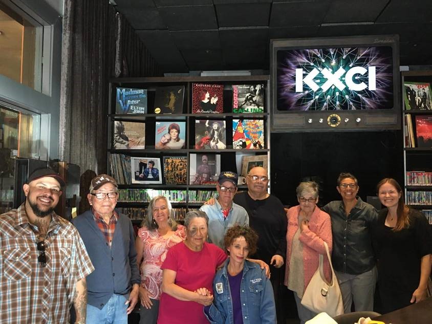 A group portrait in front of a sign that says "KXCI" and a wall of records