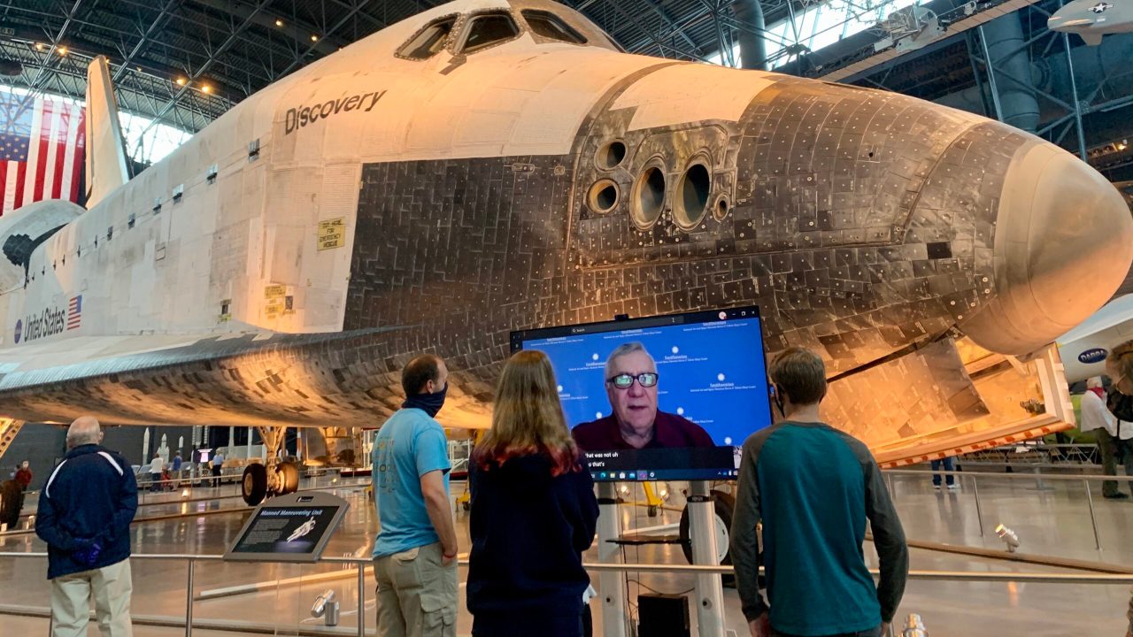 A group of visitors wearing masks speaking to a person via webcam on a TV screen in front of a plane artifact