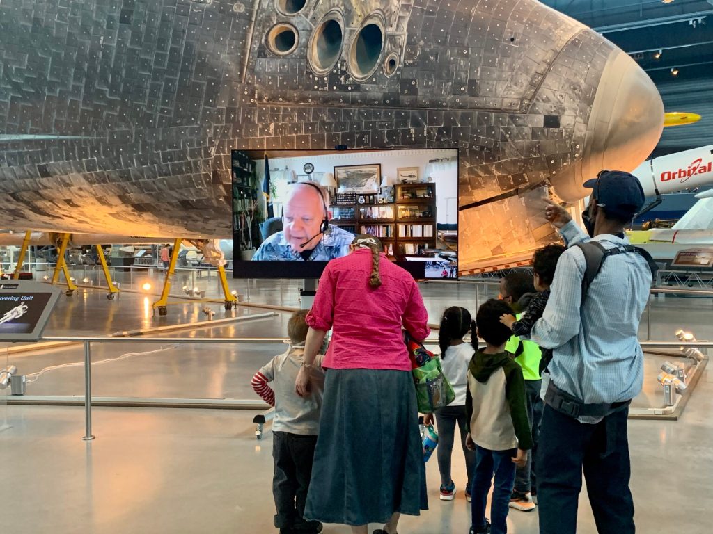 Adults with children in front of a video screen with a docent explaining an artifact plane in the background