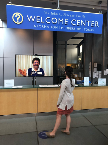 A virtual volunteering waving at a masked visitor from a video screen behind a desk labeled "The John L. Plueger Family Welcome Center"