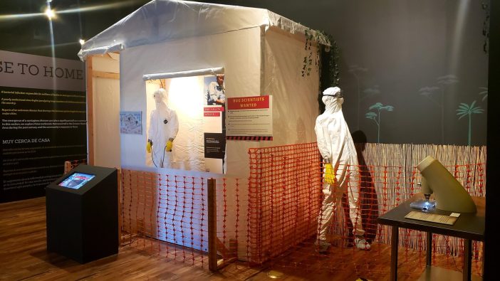 White tent in exhibition hall with outbreak panels inside and out. yellow sign in distance reads Bug Off. a screen with an interactive in front of tent.