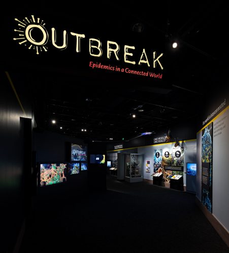 Dark lighted entrance to Outbreak exhibit with multiscreen video and displays in distance