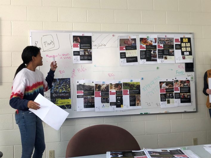 A student in a rainbow sweatshirt holding a marker pointing at a white board with printouts of panels.