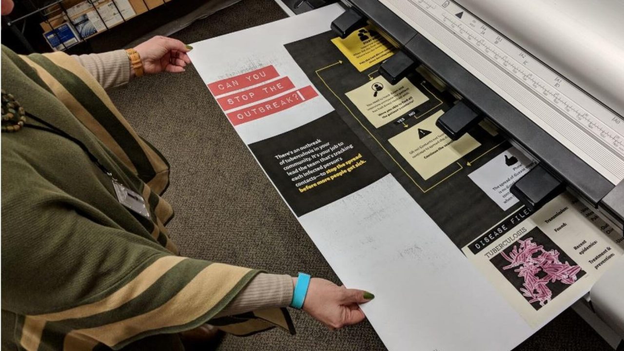 Person in green pulling large poster paper out of printer