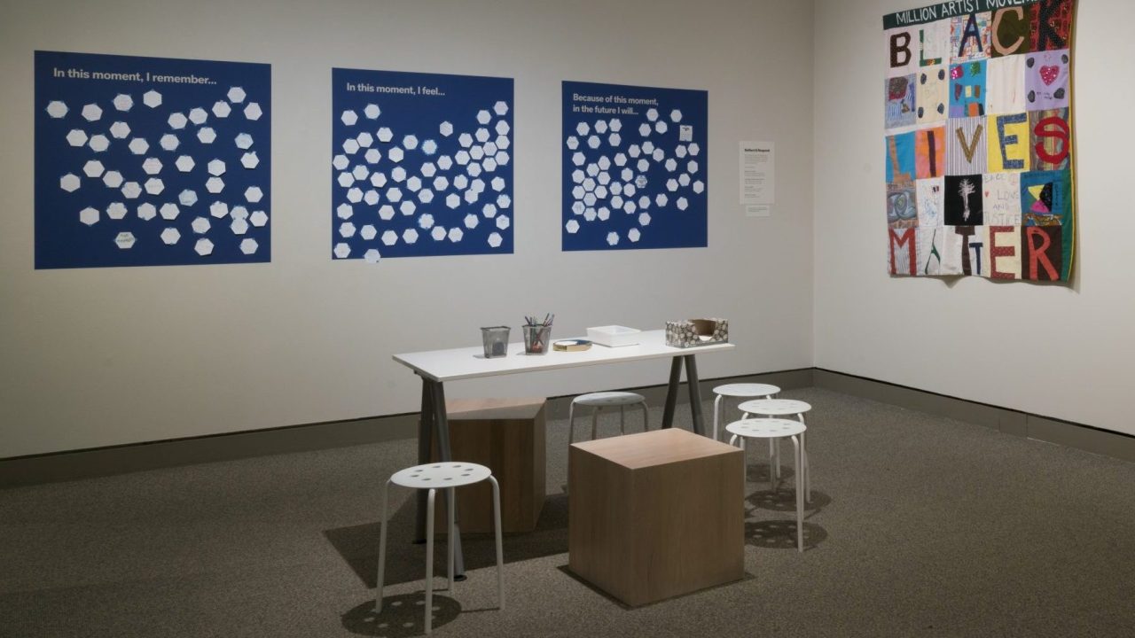 A museum gallery space with Black-Lives-Matter-themed art and a workstation for answering questions on poster boards: "In this moment, I remember...," "In this moment, I feel...," and "Because of this moment, in the future I will..."