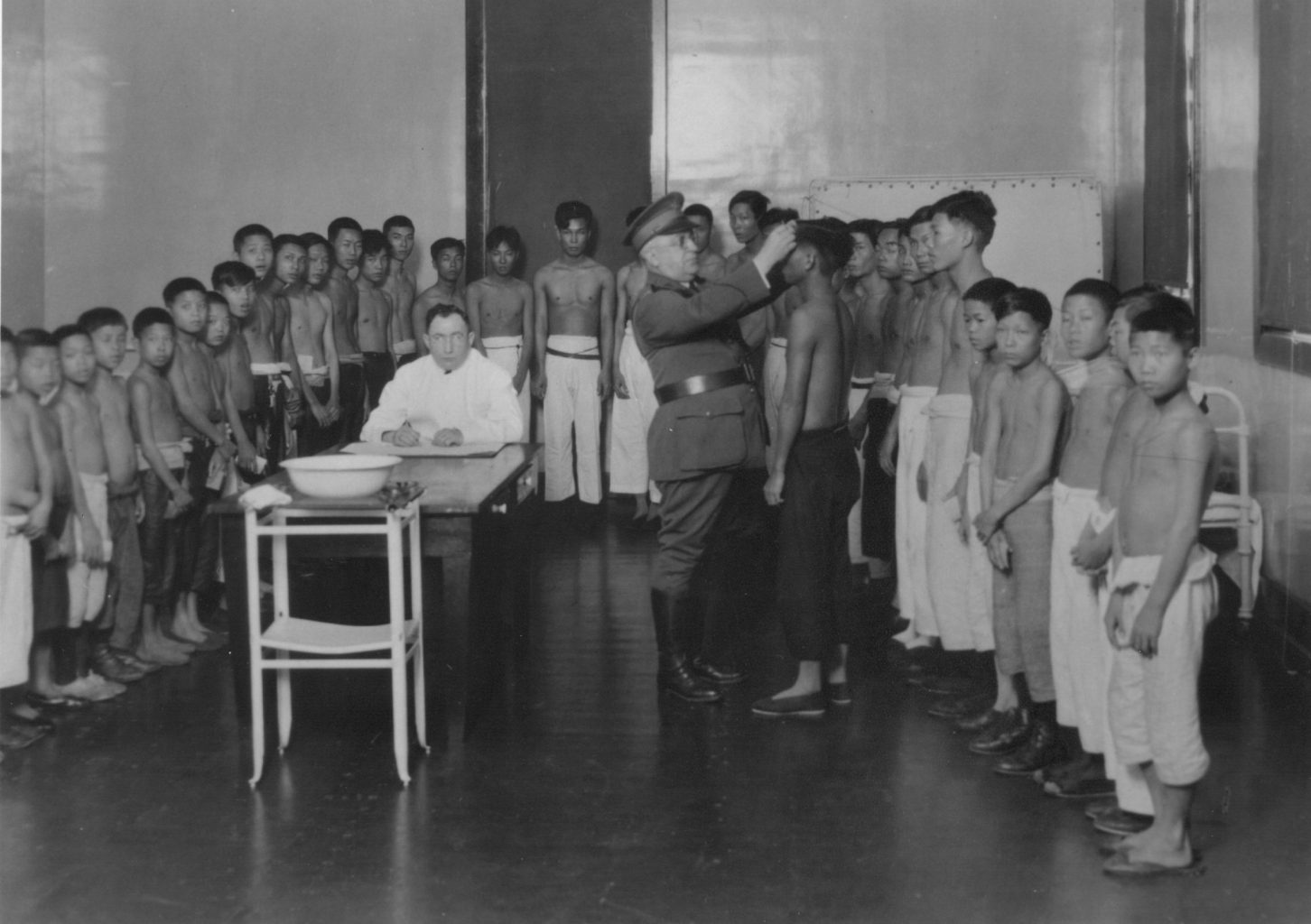 A group of Asian children and teenagers standing against the wall in a room with a white doctor and uniformed official, who is inspecting the head of one of the boys.