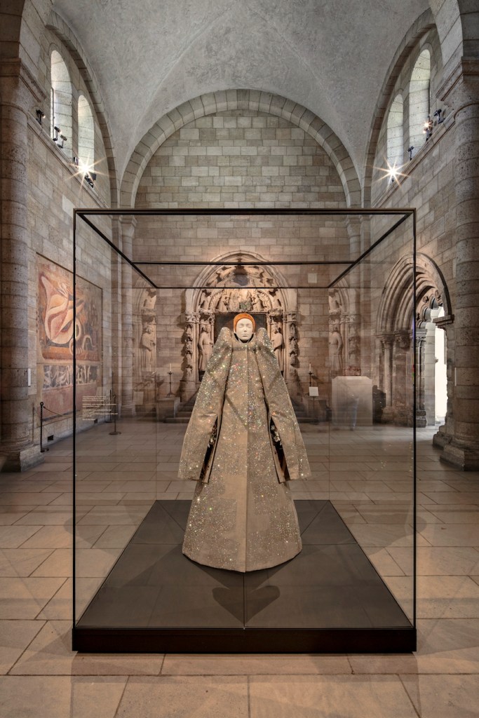 An interior view of a museum space with a mannequin wearing a garment resembling a liturgical robe inside a glass case