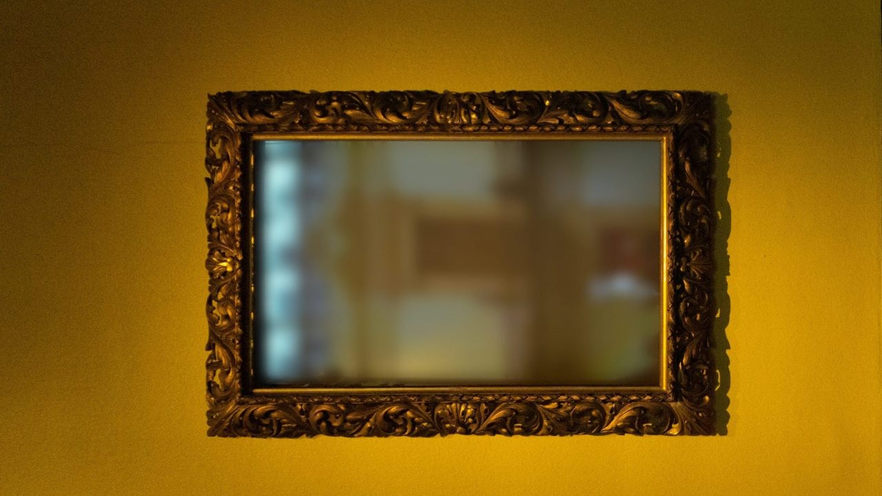 A framed mirror on a yellow gallery wall with a hazy reflection of a museum interior