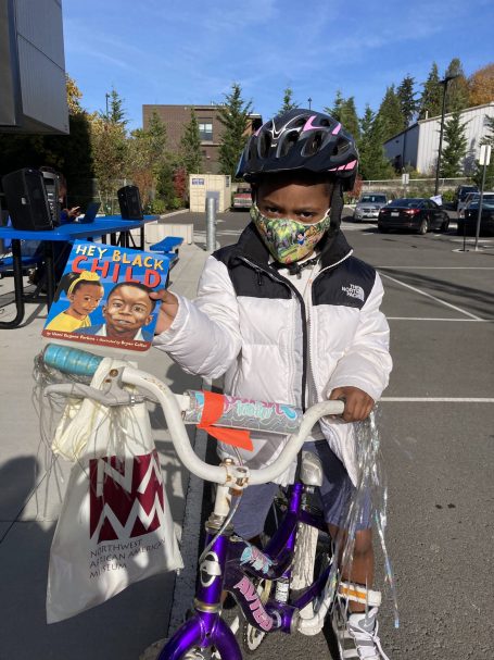 A child on a bicycle holding up a book called "Hey Black Child"